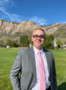 Zachary is smiling and standing in front of a blurred nature background with trees, mountains and a blue sky. He is wearing a gray suit with a white shirt and bright pink tie.