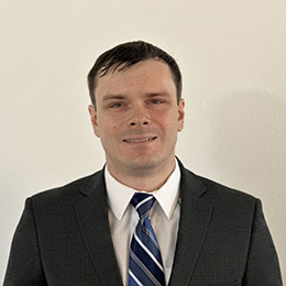 A photo of James, a man with short dark hair, wearing a dark gray suit jacket, a white dress shirt, and a blue and white striped tie. He is standing against a plain light-colored background and is looking directly at the camera with a slight smile.