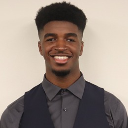 A photo of Joshua, an African American man who is smiling at the camera. He is wearing a navy-blue vest, with a gray dress shirt underneath. He has black hair which is faded on the sides with a well-kept longer top.