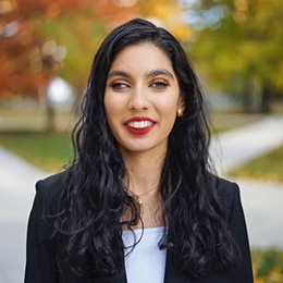 A photo of Manahil smiling at the camera. She is a Pakistani female with a light skin tone, long wavy dark hair, green eyes, and wearing a white top and a navy blue blazer.