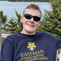 A photo of Noah, a white male with  short, light-colored hair and black sunglasses, smiling broadly at the camera. He wears a navy blue t-shirt with yellow text that reads "EASTMAN SCHOOL OF MUSIC, UNIVERSITY of ROCHESTER" along with a logo featuring the word "MELIORA" above it. The ocean is visible in the background as is a forested shoreline in the distance.