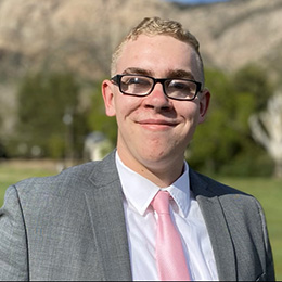 A photo of Zachary, an individual with light hair and wearing glasses, smiling. HE is wearing a gray suit coat with a white shirt and pink tie. There is also a background with green trees and brown mountains with a blue sky.