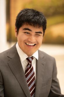 A photo of a smiling Nikhil Dadlani. He wears a gray suit jacket, white dress shirt, and a striped tie in burgundy and blue colors.