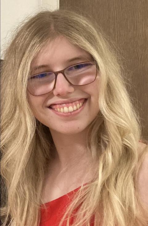 A photo of Kaleigh, a light-skinned woman with wavy blonde hair and glasses, wearing a red dress and smiling at the camera 