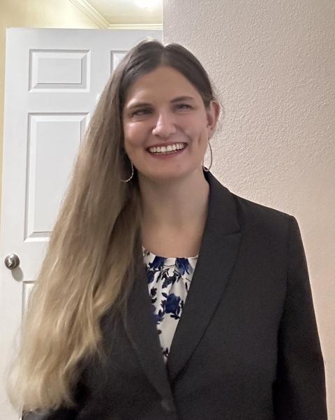 Picture of Katherine from the waist up, standing indoors against a white background and smiling confidently at the camera. She wears a charcoal blazer over a blue-and-white floral blouse, and her jewelry consists of thin, silver hoop earrings. She has dimples and long, light-brown hair. 