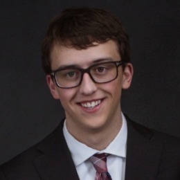 A photo of Logan smiling with medium length dark brown hair, dark brown eyes, white skin, glasses, and a black suit coat over a white dress shirt with a red tie against a dark grey background