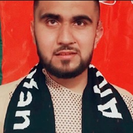 A photo of Ammar wearing a beige parahan tunban, which is traditional Afghan clothing. He has a shawl around his neck decorated with an Afghanistan flag. Behind him is the black, green and red Afghanistan flag pinned to the wall.