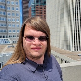 A photo of Kyle outside in the sun with downtown buildings behind him. He is wearing a dark grey polo shirt and wire frame sunglasses. Kyle has a light medium skin tone and short full facial hair with long dark blonde hair falling mostly behind his shoulders.