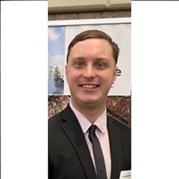 A photo of Hunter, smiling at the camera in a black suit with a dusty, pink dress shirt and a black tie. Hunter is a Caucasian male. Hunter has blue eyes and brown hair combed to the side.
