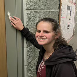 A photo of Lauren, a woman with white skin and light brown hair that is pulled back, is smiling and looking at the camera. She’s wearing a black sweatshirt unzipped over a maroon shirt with a white embroidered design and a silver chain necklace. She is standing next to the door to her old dorm room, reaching up to touch the small white mezuzah that hangs on the doorframe. 