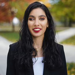 A photo of Manahil smiling at the camera. She is a Pakistani female with a light skin tone, long wavy dark hair, green eyes, and wearing a white top and a navy blue blazer.