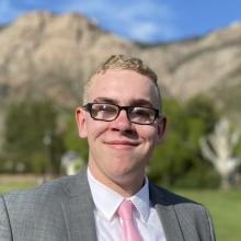 Zachary, a young man wearing a gray suit and pink tie, smiles at the camera while standing in a grassy field. Behind him, dramatic rocky mountains rise beneath a bright blue sky.