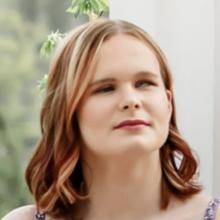 Stormy, a young woman with auburn hair, smiles softly at the camera. She wears light makeup, and her hair falls loose around her shoulders.