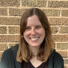 Elizabeth, a young woman with light skin and shoulder-length brown hair, stands in front of a textured brick wall. Her hair is parted slightly off-center and falls in soft waves around her face. She wears a black V-neck top and a chunky-knit dark green cardigan.