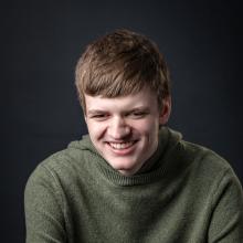 Benjamin, a young man with brown hair, sits against a gray background wearing a green shirt and smiling at the camera.