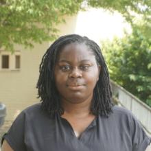 Soso, a young Black woman with shoulder-length locks, stands outdoors in front of green trees and a building. She wears a dark top and faces the camera.