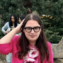 Noor, a young woman with fair skin, long brown hair, and black-framed glasses, walks toward the camera wearing a bright pink pantsuit and a white-and-pink printed top. She carries a small white handbag.