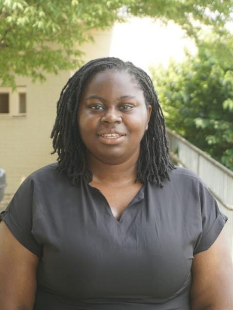 Osolu is wearing a black blouse and she is standing outside  in an area with  trees behind her.  
