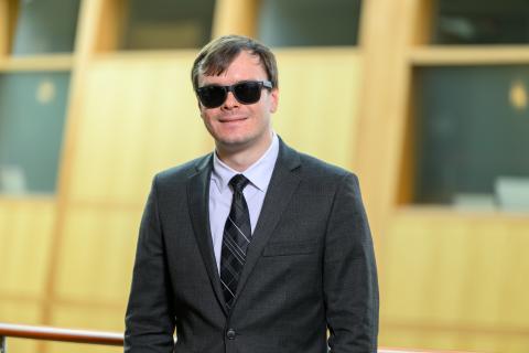 Waist‑up photo of a man in black sunglasses, charcoal‑gray suit, white dress shirt, and a diagonal‑striped tie, smiling as he stands by a railing inside a modern building with warm wood paneling and frosted glass in the background.