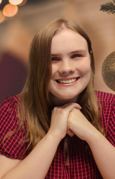 A young blond woman smiling at the camera. She is wearing a red and black dress with her hands crossed under her chin.