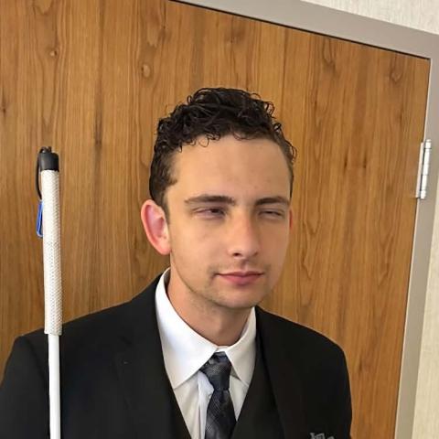 Bryson, a young man dressed in a black suit with a vest, tie, and pocket square, stands indoors in front of a wooden door. He holds a long white cane in his right hand and rests his left hand in his pocket.